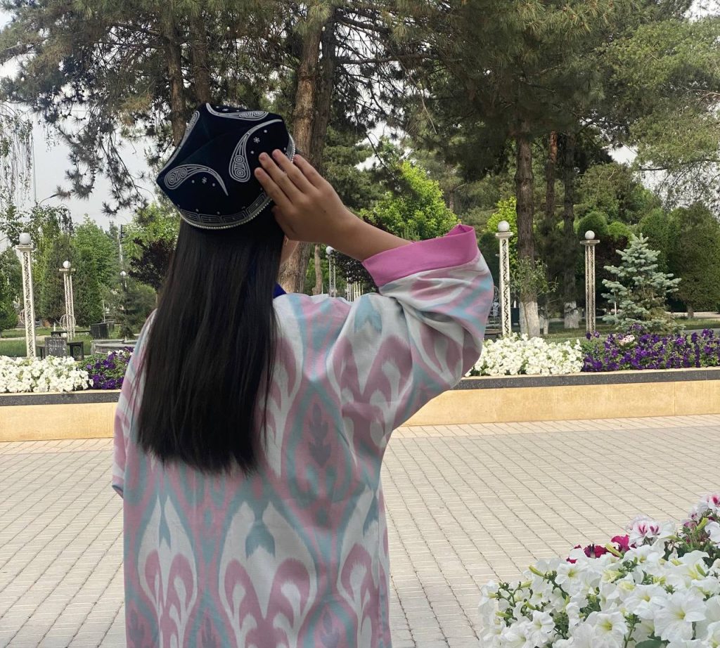 Young Central Asian teen girl with long dark hair, a black and white cap, and a patterned green and pink and white coat. She's outside by a concrete path and petunias.