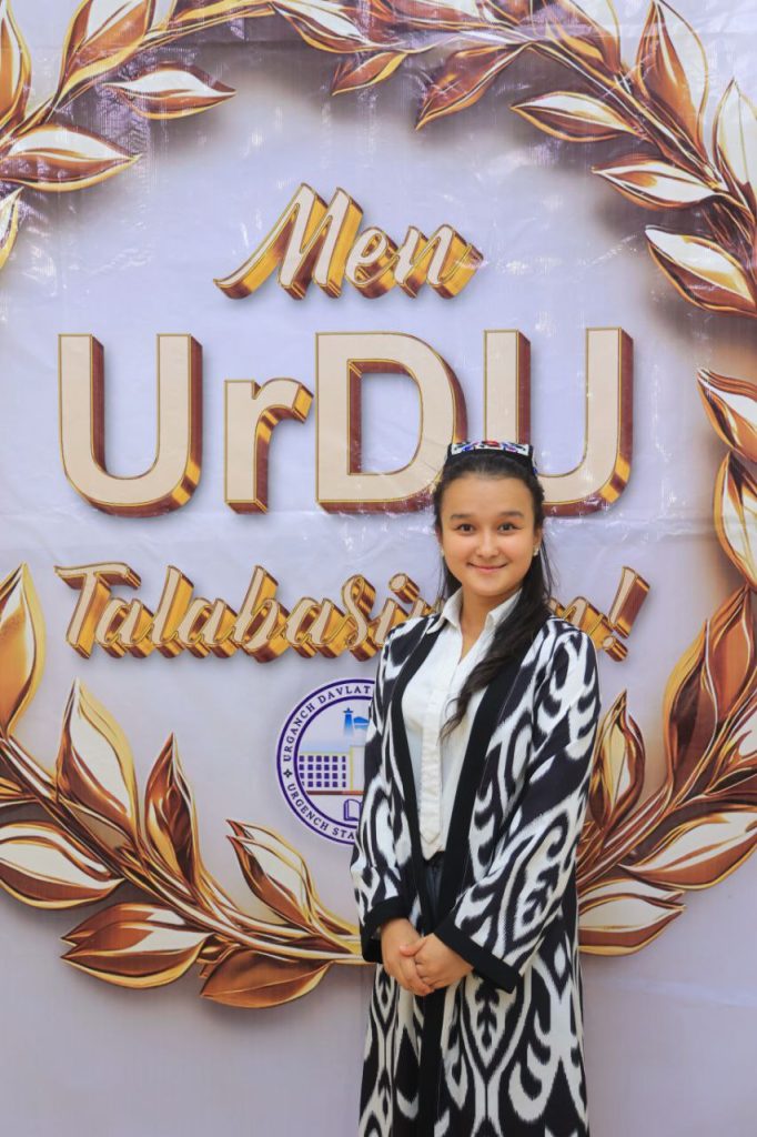 Central Asian teen girl with long dark hair and an embroidered headdress in a long black and white coat over a white collared shirt standing on stage in front of a signboard with gold letters. 