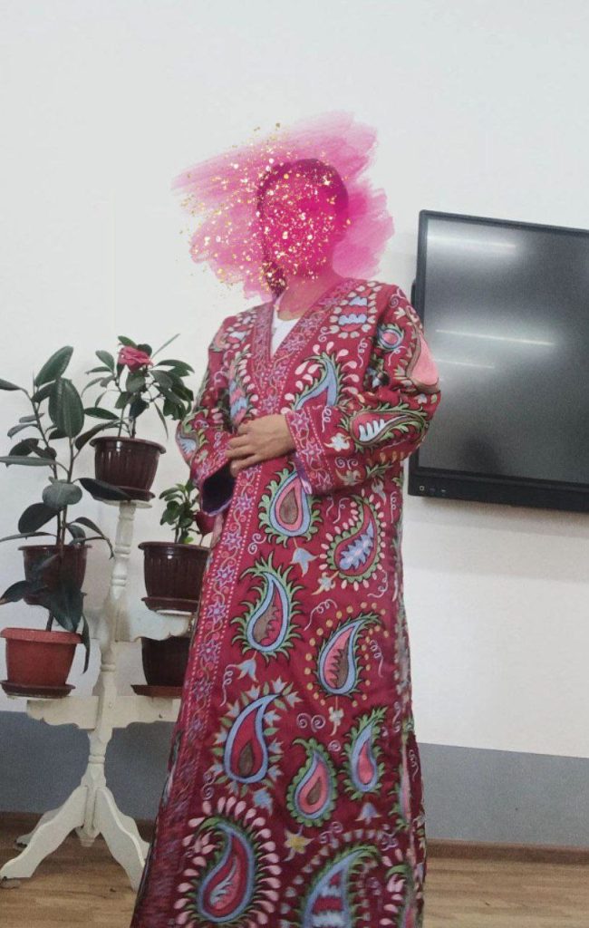 Young Central Asian woman with a patterned pink and green coat standing next to a TV screen and a set of leafy houseplants. 