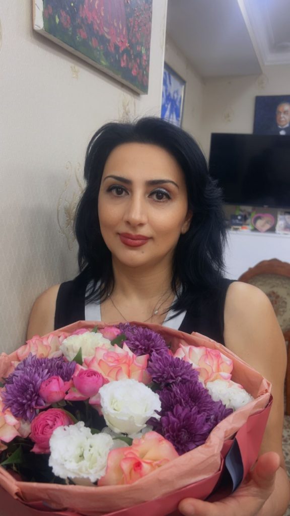 Central Asian woman with short dark hair and a black sleeveless top holding a bouquet of pink and purple flowers. 
