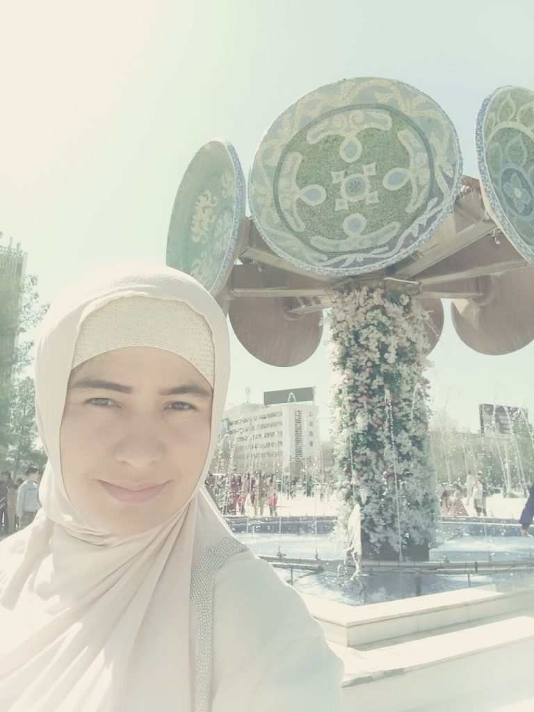 Young Central Asian woman in a light tan headscarf and top, in front of a public statue with flowers, a pond, and circular bowl art work.
