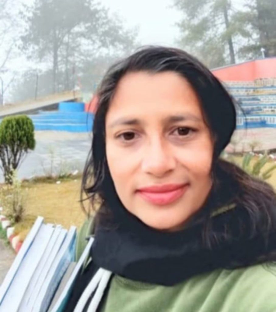 South Asian young woman with short dark hair and a green sweater and black scarf outside in front of a blue staircase and green grass and bushes. 