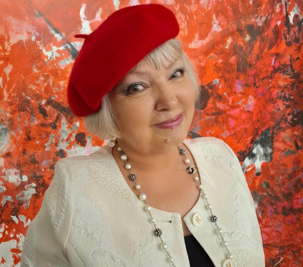 Middle aged light skinned woman with brown eyes and short blonde hair in a red beanie and pearl necklace and white top with a white and orange background. 