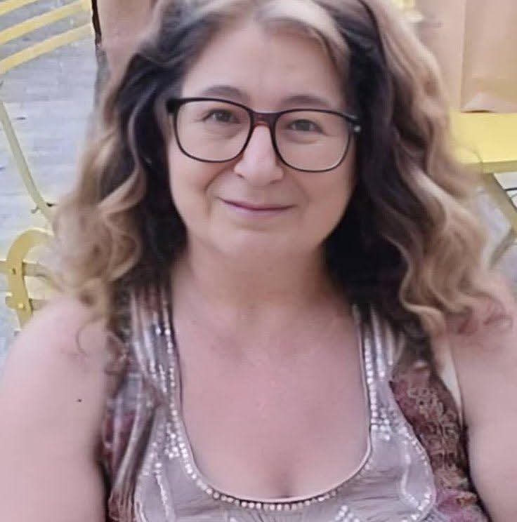 Middle aged European woman with curly brown hair, glasses, and a sleeveless top.