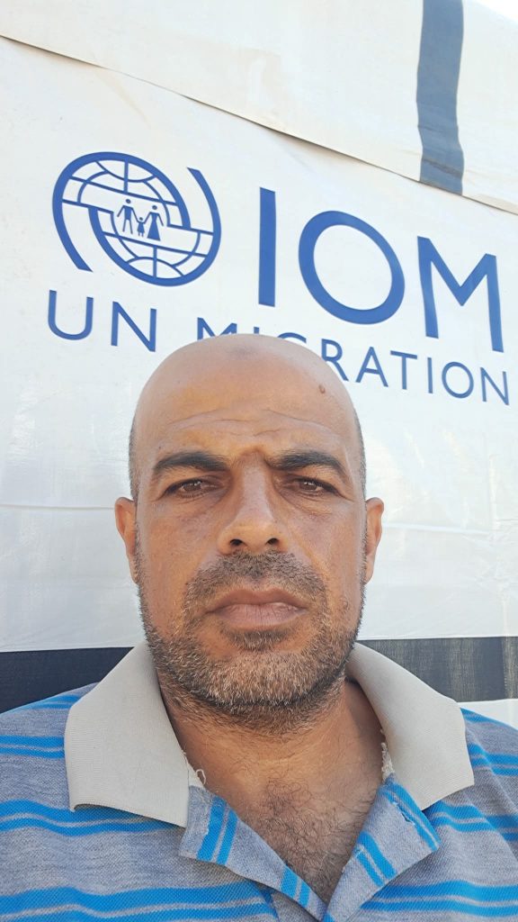 Bald Middle Eastern man, middle aged, standing in front of a sign and in a gray and white collared shirt.