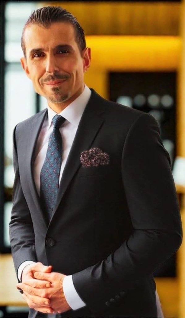 Middle aged Middle Eastern man with a trimmed mustache and beard and black suit coat and white collared shirt and blue tie. 