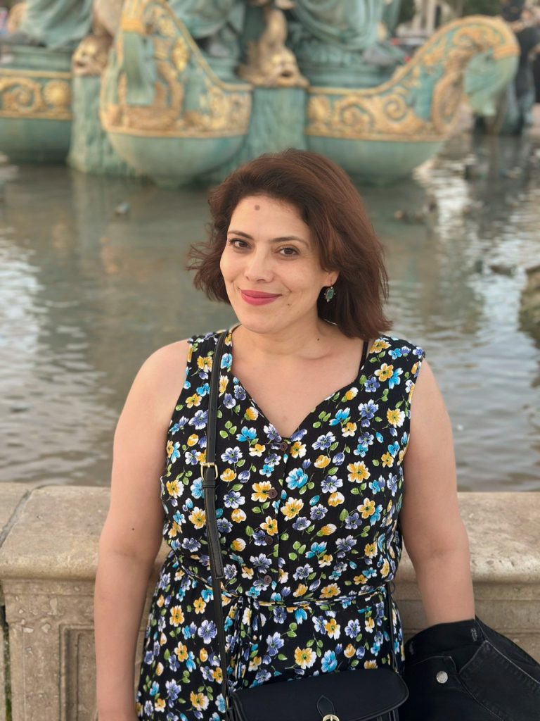 Light skinned Arab-European woman with short brown hair and a flowered dress and black purse stands in front of a pond with decorative concrete figures.