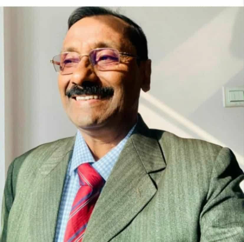 Older South Asian man with short dark hair, reading glasses, trimmed mustache, and a gray coat over a blue and white collared shirt and red tie.