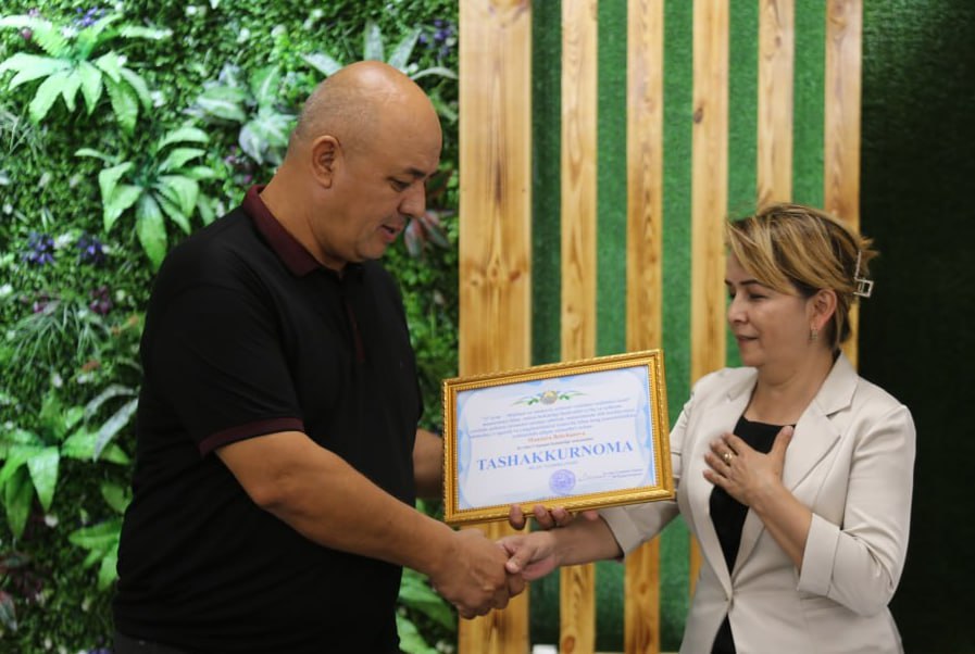 Young middle aged Central Asian woman with short brown hair up in a bun in a light colored coat over a black top receiving a certificate from a middle aged man with a bald head in a black tee shirt. They're shaking hands on stage in front of a bush with leaves and flowers.