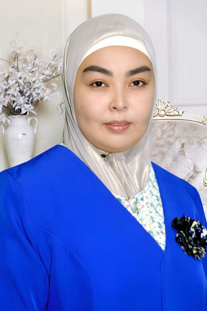 Young Central Asian woman with a white headscarf, blue coat and patterned floral undershirt, standing in front of a white cushioned chair and white flowers. 