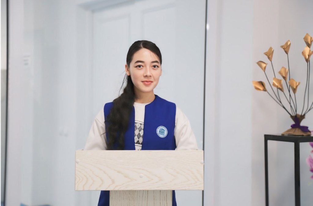 Central Asian teen girl with long dark hair and a blue vest standing at a wooden podium. 