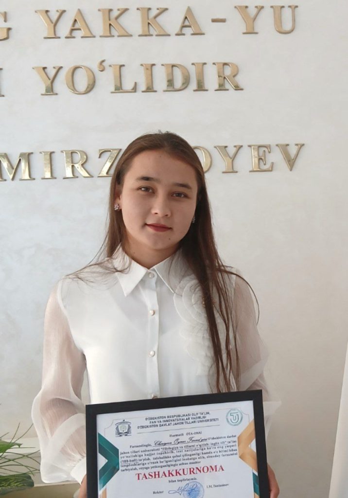 Young Central Asian woman with long dark hair and brown eyes in a white collared blouse holding a certificate. 
