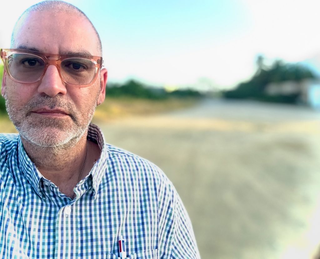 Bald middle aged light skinned man with reading glasses and a plaid collared shirt. Dry ground and trees on a hiking path in the background. 