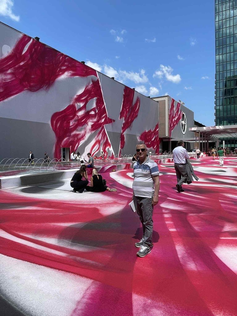 Man in jeans and a striped tee shirt standing in front of an art installation outdoors with swathes of red paint. 