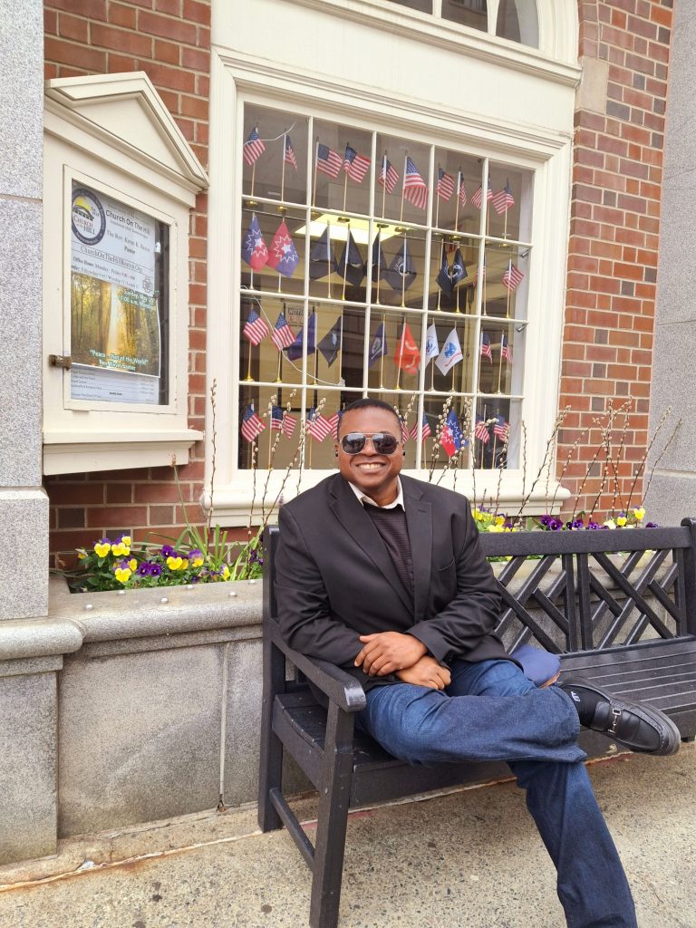 Middle aged Black man in a black coat and sunglasses seated in front of a brick building with white windows. Pansies and flags decorate the house. 