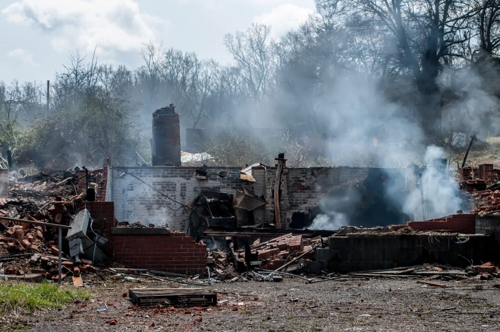 Burned out wood and brick building still steaming with trees and dirt and green grass. 
