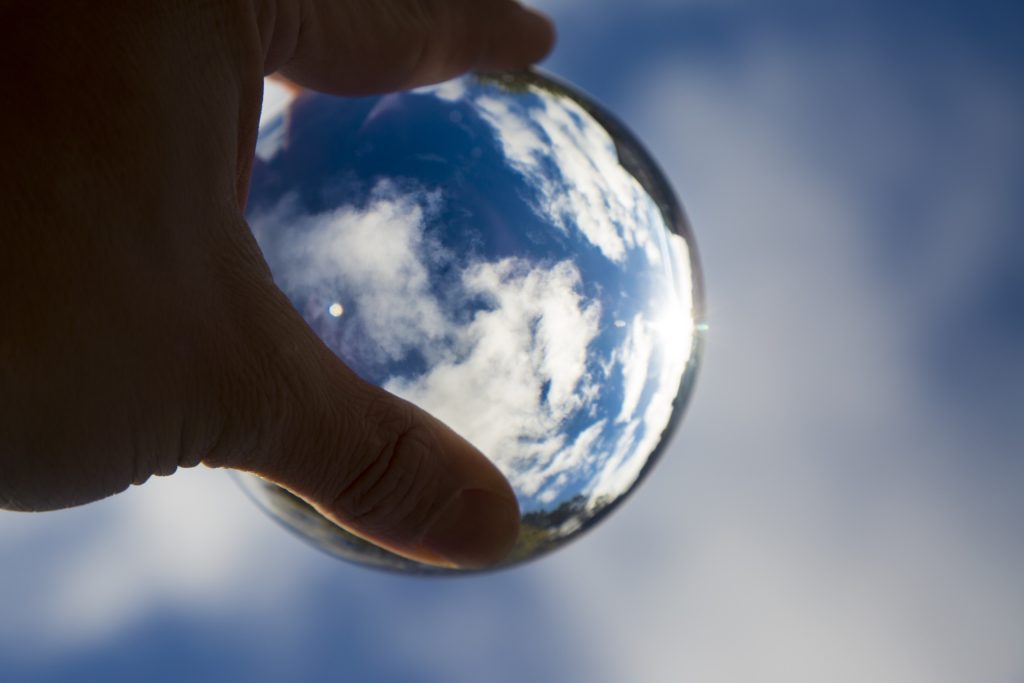 Person's hand holding a white ball and magnifying a few clouds against the sky. 