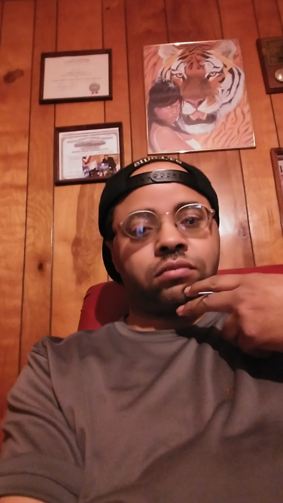 Young Black man with reading glasses in a baseball cap in a dark tee shirt seated in a chair.