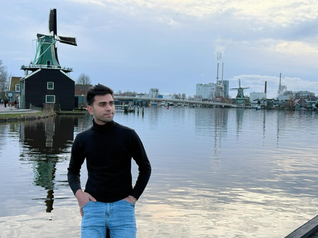 Young Rohingya (Myanmar) Muslim guy stands in front of a lake and windmill on a cloudy day. He's in a black turtleneck and blue jeans. 