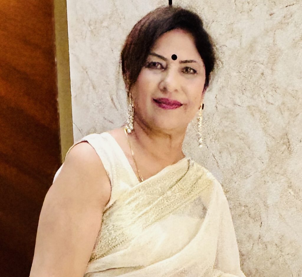 Middle aged South Asian woman with thick dark hair up in a bun, long earrings, and a tan saree.