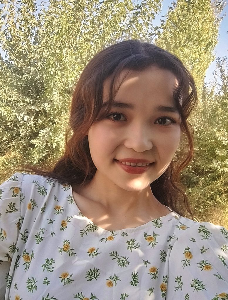 Young Central Asian woman with a floral blouse and brown curly hair in front of a green leafy tree on a sunny day.