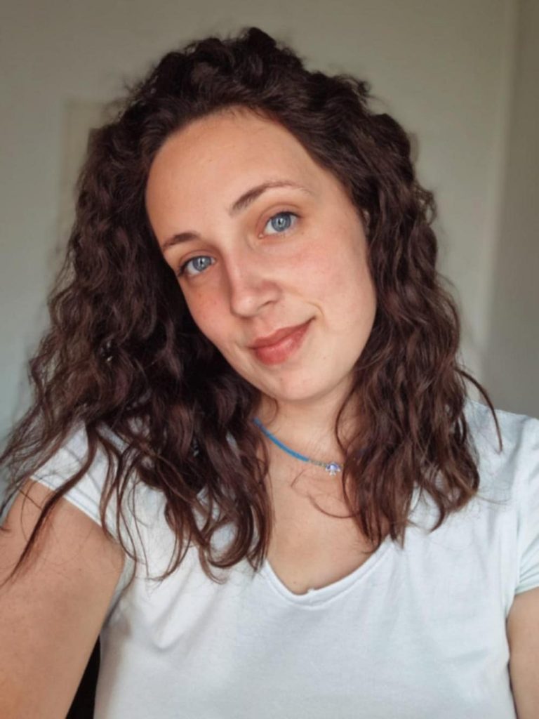 Light skinned European woman with curly brown hair, blue eyes, and a blue necklace and white tee shirt.