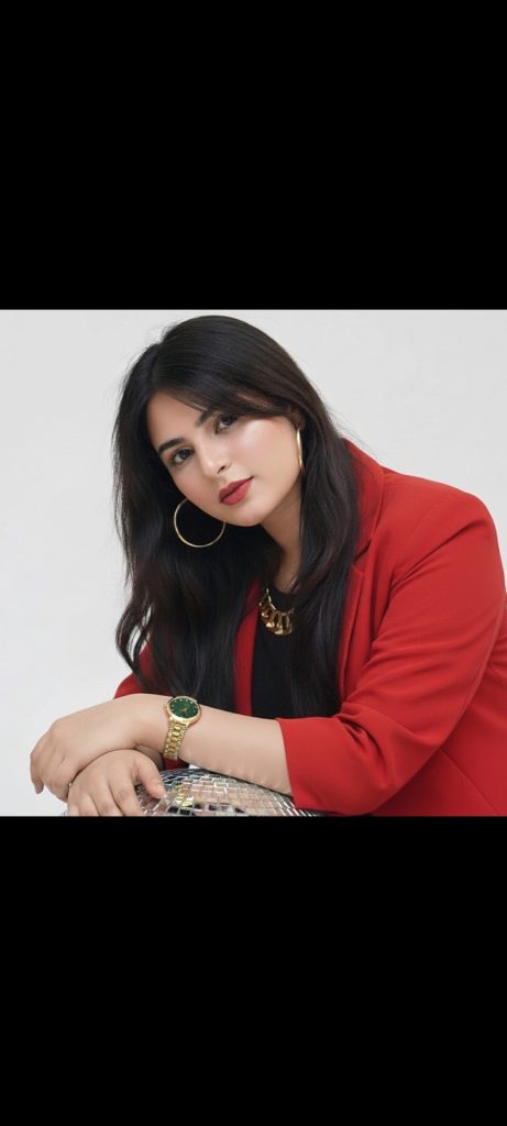 South Asian woman with long dark hair and hoop earrings and a red coat over a black blouse and gold necklace.