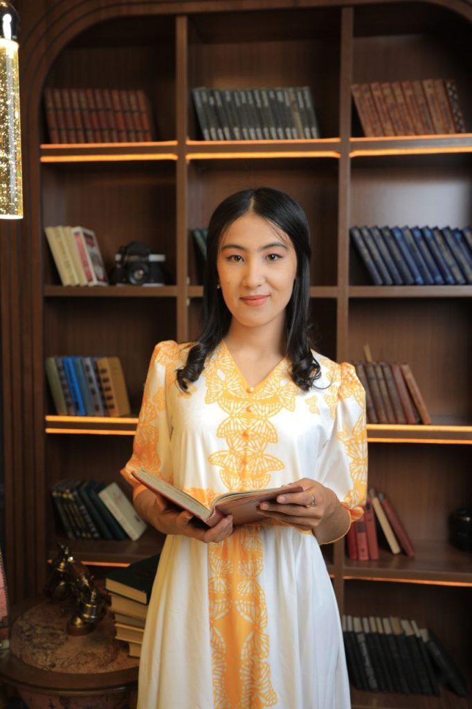 Young Central Asian woman with long dark hair and a white dress embroidered with orange, standing in front of a bookshelf.