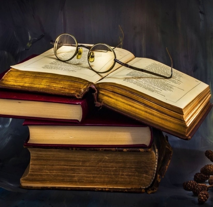 Older books stacked on top one another. The top book is open to what looks like poetry and reading glasses are on top of it.