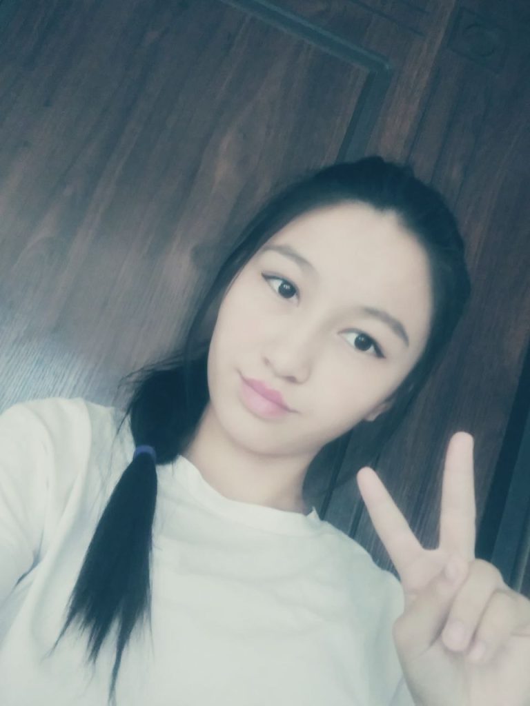 Young Central Asian woman with dark hair in a ponytail and a white tee shirt against a wood background. 