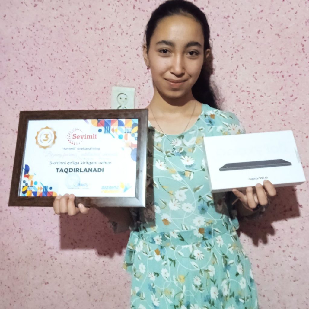 Central Asian teen girl with long dark hair in a ponytail and a green floral blouse holding up certificates.