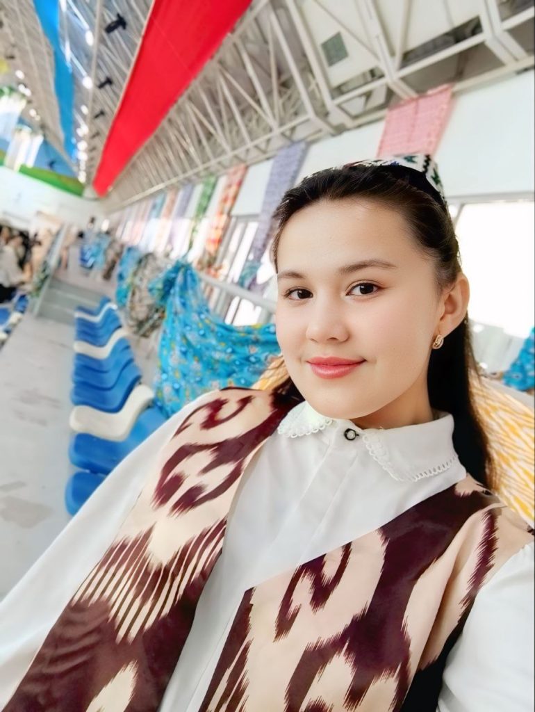 Young Central Asian woman in a large stadium with colorful banners and an embroidered cap, small earrings, long dark hair, a brown and white vest, and white blouse.