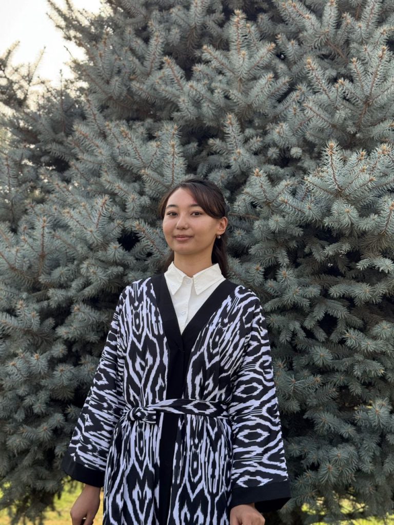 Young Central Asian woman standing in front of an evergreen tree. She's got long dark hair in a ponytail and a patterned black and white coat and white collared shirt.