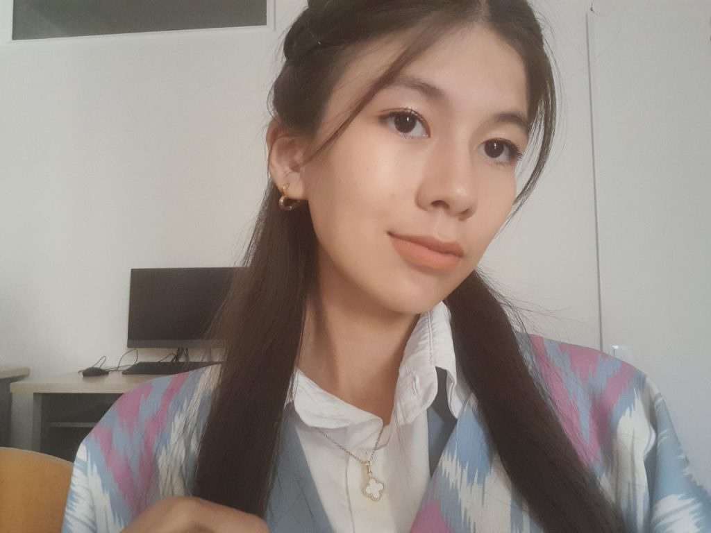 Central Asian young woman with long dark hair and brown eyes in a pink and blue and white vest and white collared shirt.