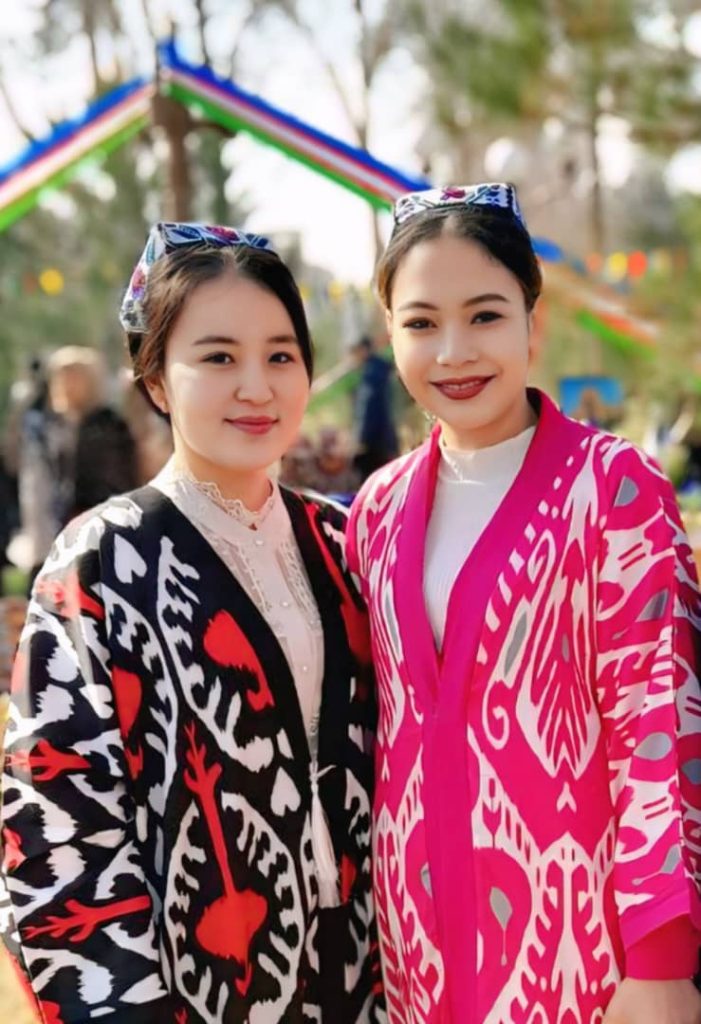 Two Central Asian young women with colorful pink and red and black light coats and embroidered headdresses standing outside at some sort of outdoor park or festival.