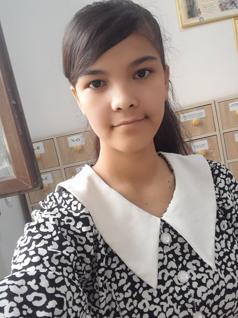 Young Central Asian teen girl with long dark hair, brown eyes, and a black and white patterned top standing in front of a set of drawers. 