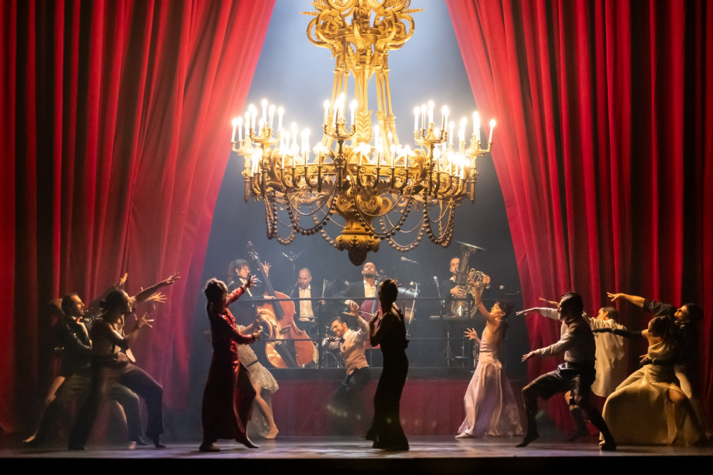 Golden chandelier above a stage with a red curtain and people in suits and ball gowns dancing in front of an orchestra with instruments. 