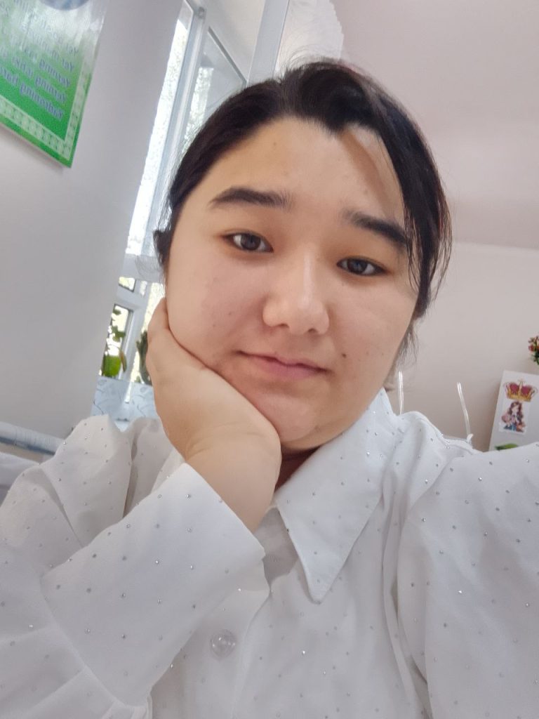 Young Central Asian woman with short dark hair and a white collared shirt, seated and resting her head on her hand. 