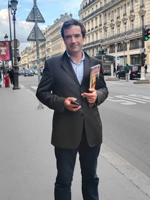 Middle aged light skinned man in a gray coat and collared shirt standing on a busy street holding a book and his phone.