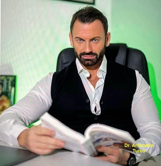 Intense looking white man, 30s-40s, trimmed hair, beard, mustache, black vest and white collared shirt, seated in a chair and reading a book.