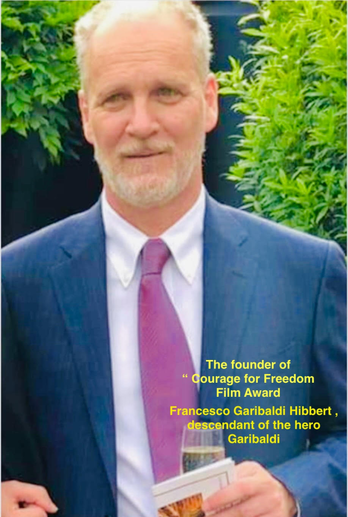 Older white man with short gray hair, a black coat, and a red tie and white collared shirt standing outdoors in front of green bushes. 