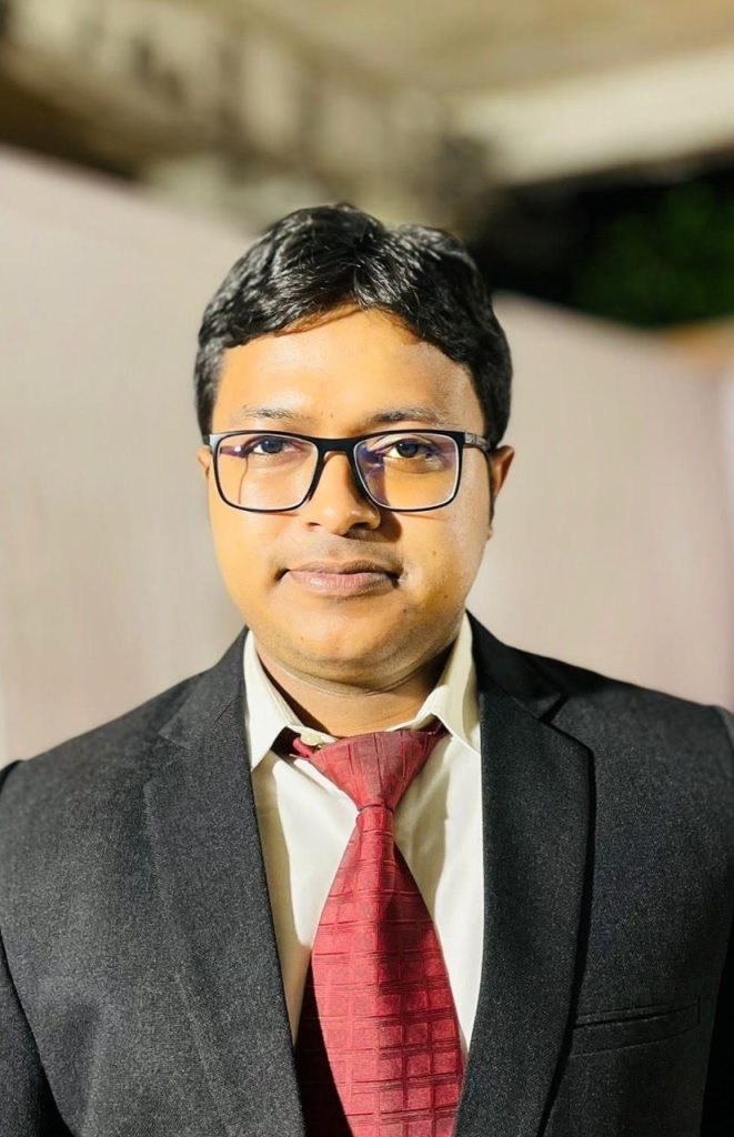 Young South Asian man with short dark hair, reading glasses, and a black suit and red tie.