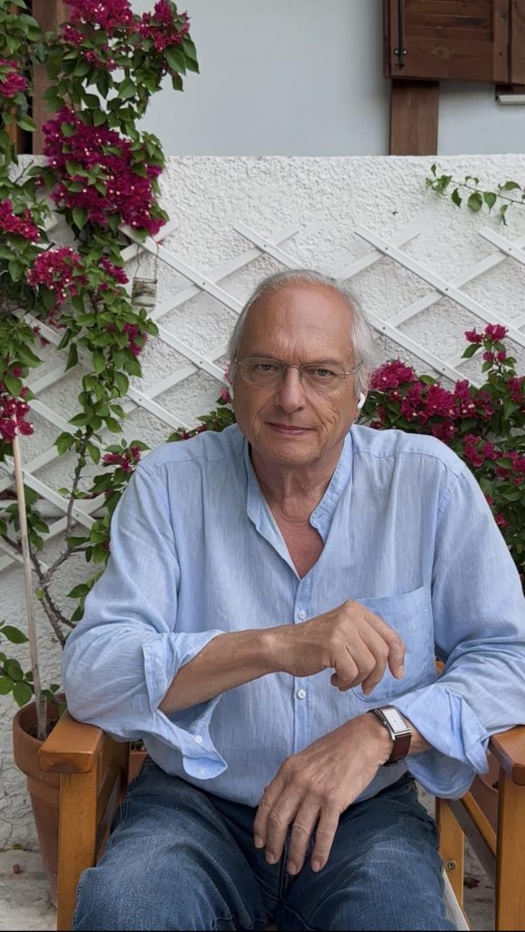 Older European light-skinned man with gray hair and reading glasses in a light blue collared shirt and vines of purple flowers.