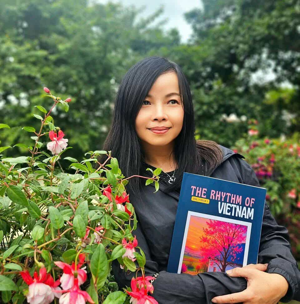 Young East Asian woman with long dark hair, brown eyes, a black coat standing in front of blooming fuschia plants. She's holding a book, The Rhythm of Vietnam.