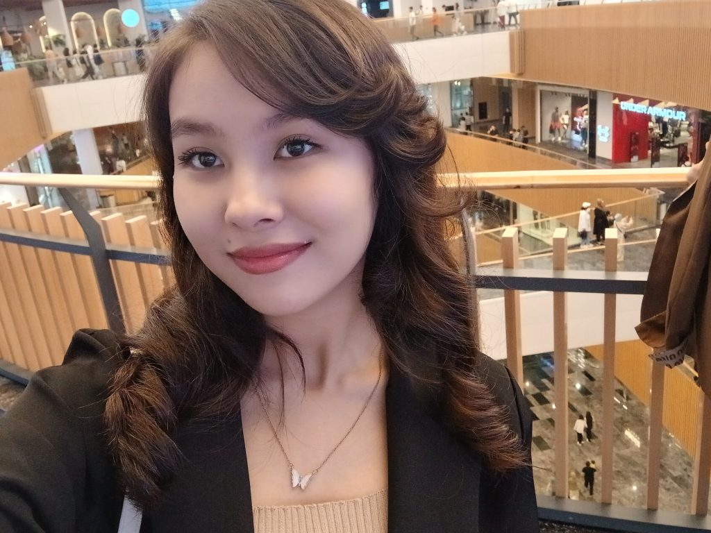 Young Central Asian woman with curly brown hair, brown eyes, and a dark top and small necklace. 