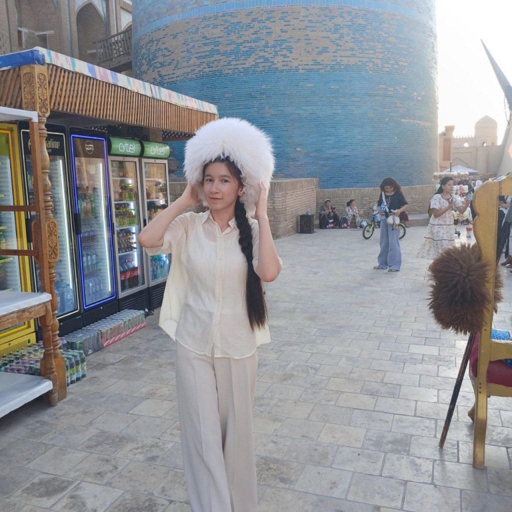 Young Central Asian woman in a white fluffy headdress and light colored top and pants outside near some light blue and brown buildings. Historical looking area.