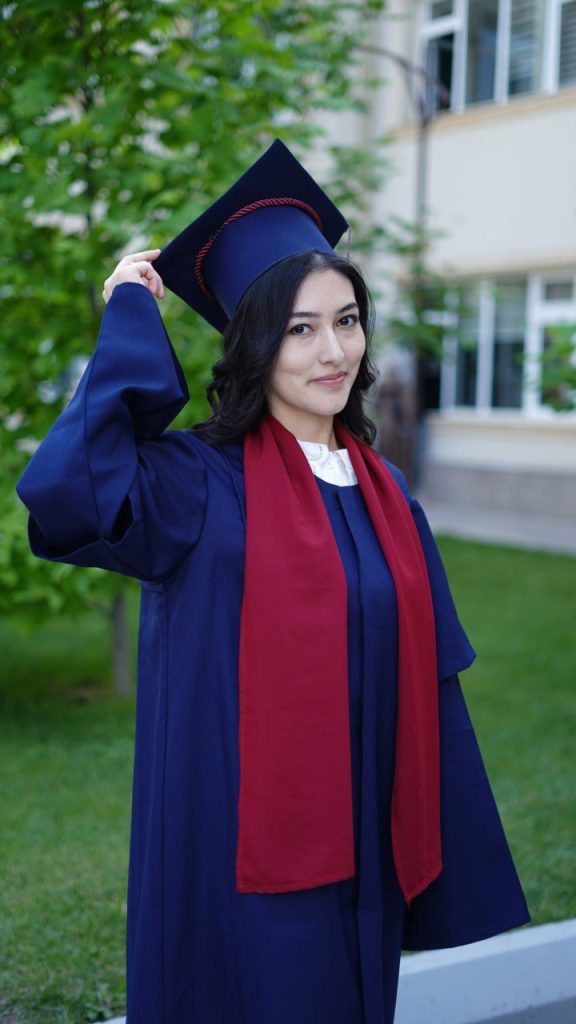 Young Central Asian woman with a blue graduation cap and gown and red sash standing outside near a green tree on a lawn near a white school building. 
