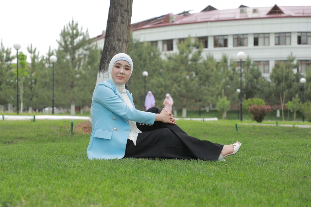 Young Central Asian woman in a blue jacket and headscarf and black pants and sandals seated on a green lawn near a tree outside a large building.