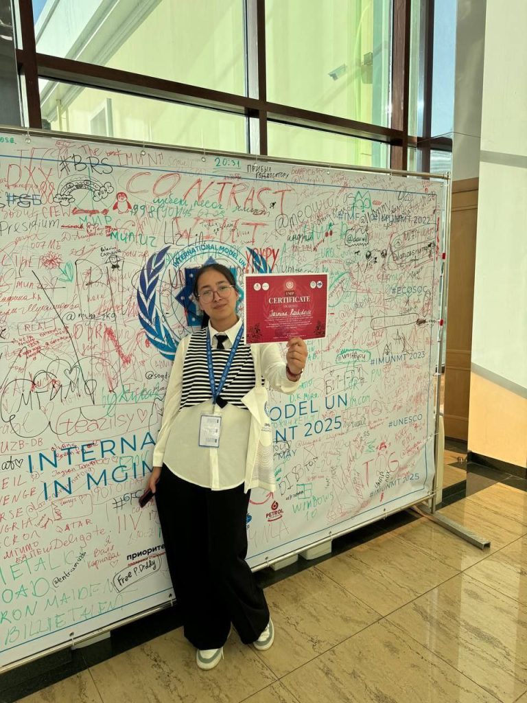Central Asian woman with long straight dark hair, reading glasses, and a white top and black pants, holding a red certificate in front of a white board with a whole group of signatures from Model UN participants. 