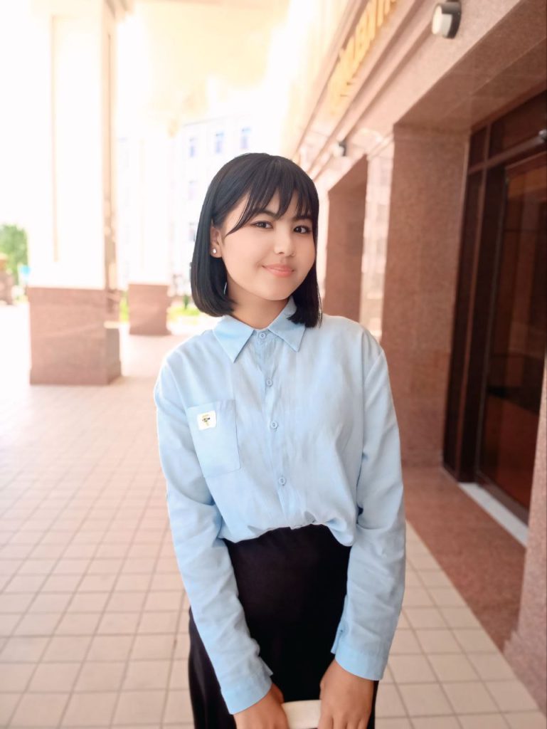 Young Central Asian teen girl with short dark hair, small pearl earrings, brown eyes, and a white collared shirt and black pants, posing outside on concrete by an open window. 